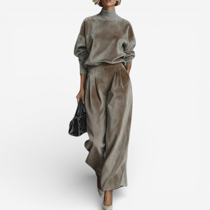 Model in taupe contemporary set with turtleneck and wide-leg pants, holding a black bag, against a white background.