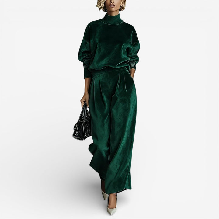 Model in emerald green contemporary set with turtleneck and wide-leg pants, carrying a black bag, on a white backdrop.