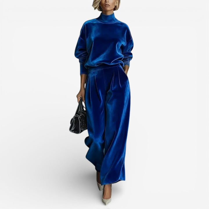 Model in royal blue contemporary set with turtleneck and wide-leg pants, holding a black bag, standing against white.
