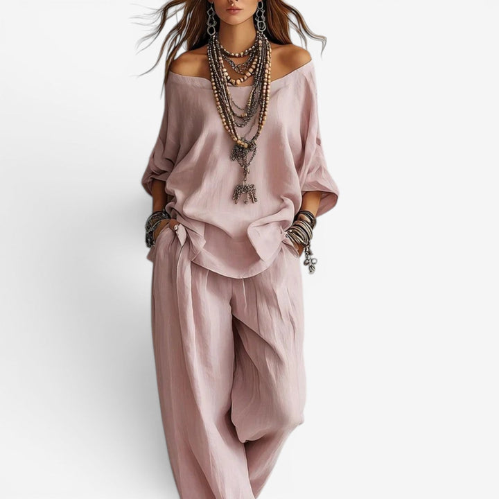 Elegant set in soft pink with off-shoulder top, relaxed pants, layered necklaces, and bracelets. Studio background.