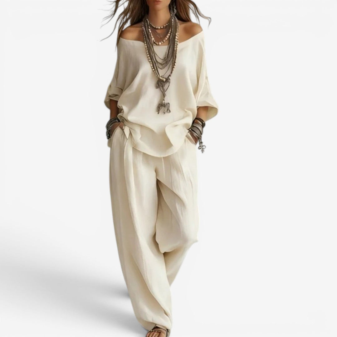 Elegant set in ivory with off-shoulder top, loose pants, layered necklaces, and bracelets. Studio background.