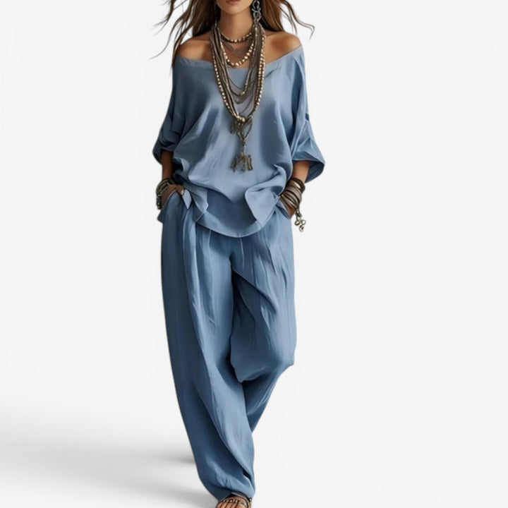 Elegant set in blue featuring off-shoulder top, flowing pants, layered jewelry. Studio background.