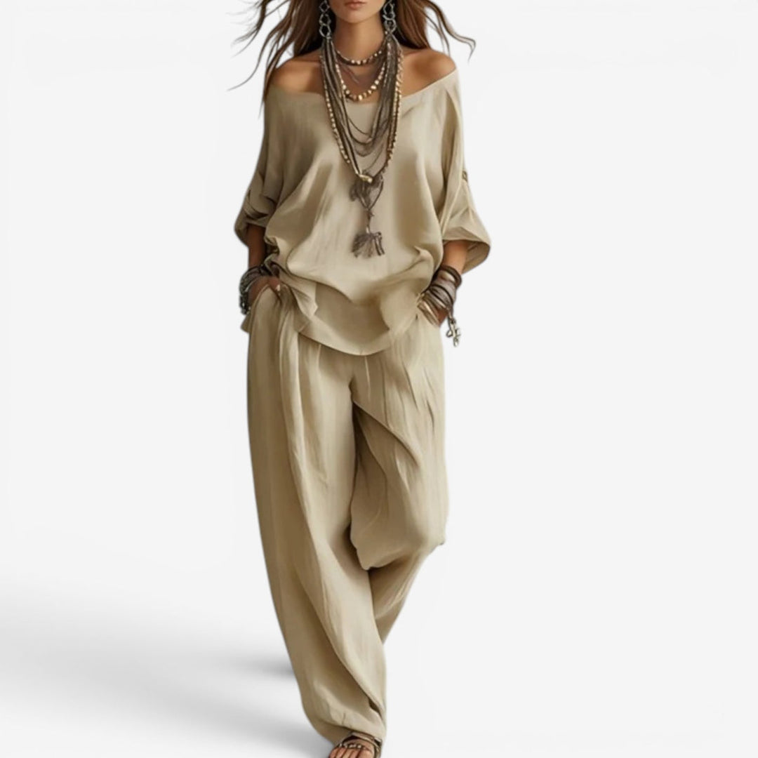 Elegant set in beige with off-shoulder top, wide pants, layered necklaces, and bracelets. Studio background.