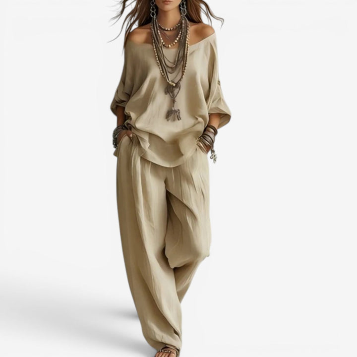 Elegant set in beige with off-shoulder top, wide pants, layered necklaces, and bracelets. Studio background.