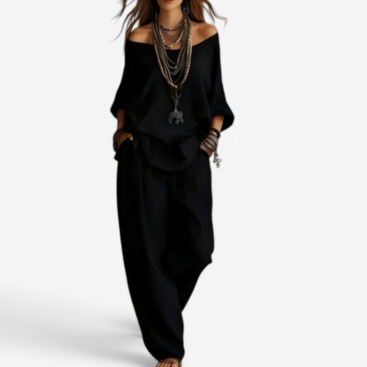 Elegant set in black featuring off-shoulder top, relaxed pants, and layered jewelry. Studio background.