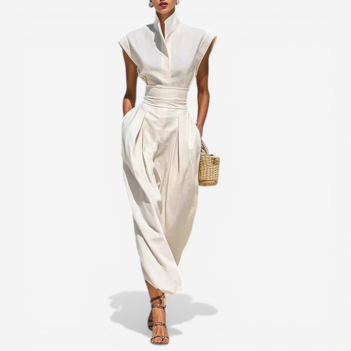 Model in elegant cream jumpsuit with high collar and wide waistband, holding a wicker purse against a white background.