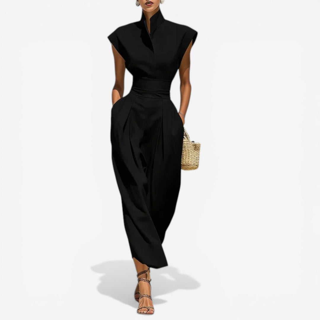 Model in elegant black jumpsuit with high collar and wide waistband, holding a wicker purse against a white background.