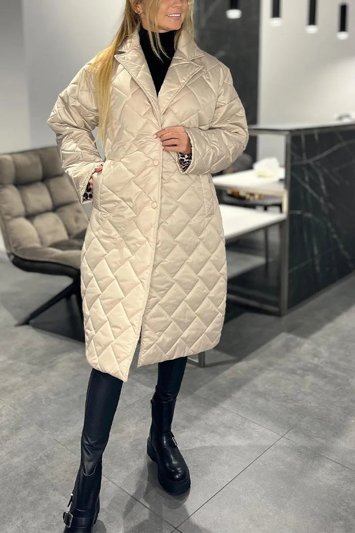 Model in a light beige quilted coat, standing in a modern indoor space with gray floors and a cushioned chair.