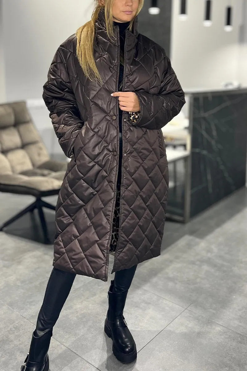 Model wearing a dark brown quilted coat, standing in a modern indoor setting with gray flooring and a cushioned chair in the background.