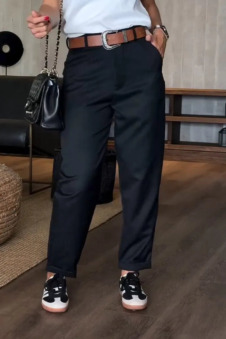 Black tailored pants indoors, styled with a brown belt, and paired with sneakers and a black handbag.