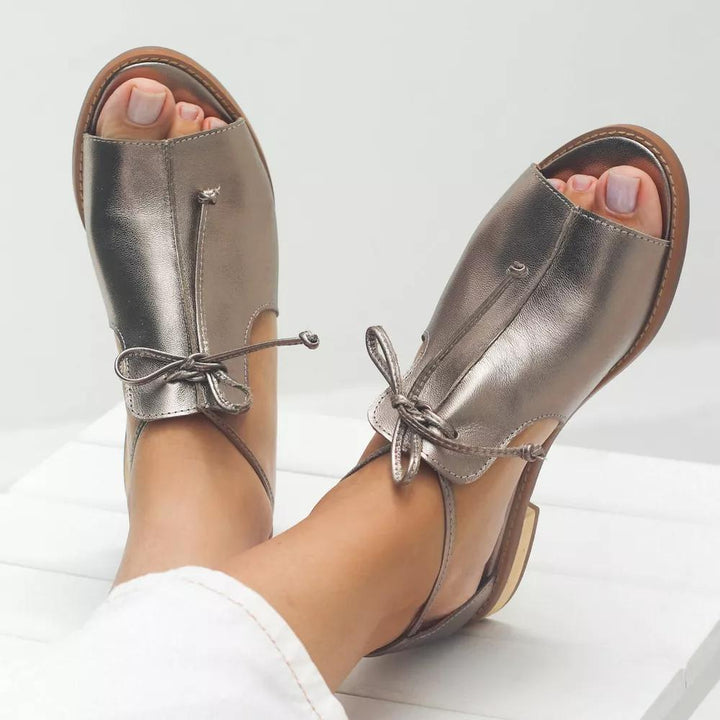 Metallic leather sandals with bow detail on feet, indoor white setting.
