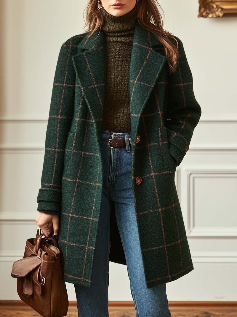 Model wearing a green plaid tweed coat over a green turtleneck sweater with blue jeans, holding a brown handbag in an elegant interior setting.