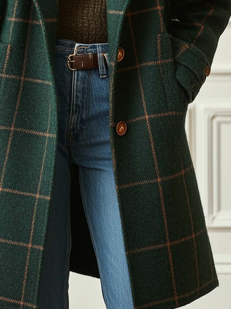 Close-up of a green plaid tweed coat with button details, worn over blue jeans, set against an indoor backdrop.