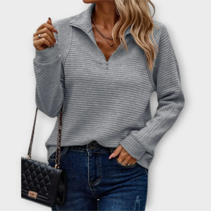 Woman wearing a grey ribbed half-zip pullover, holding a black quilted purse, paired with jeans, on a plain backdrop.
