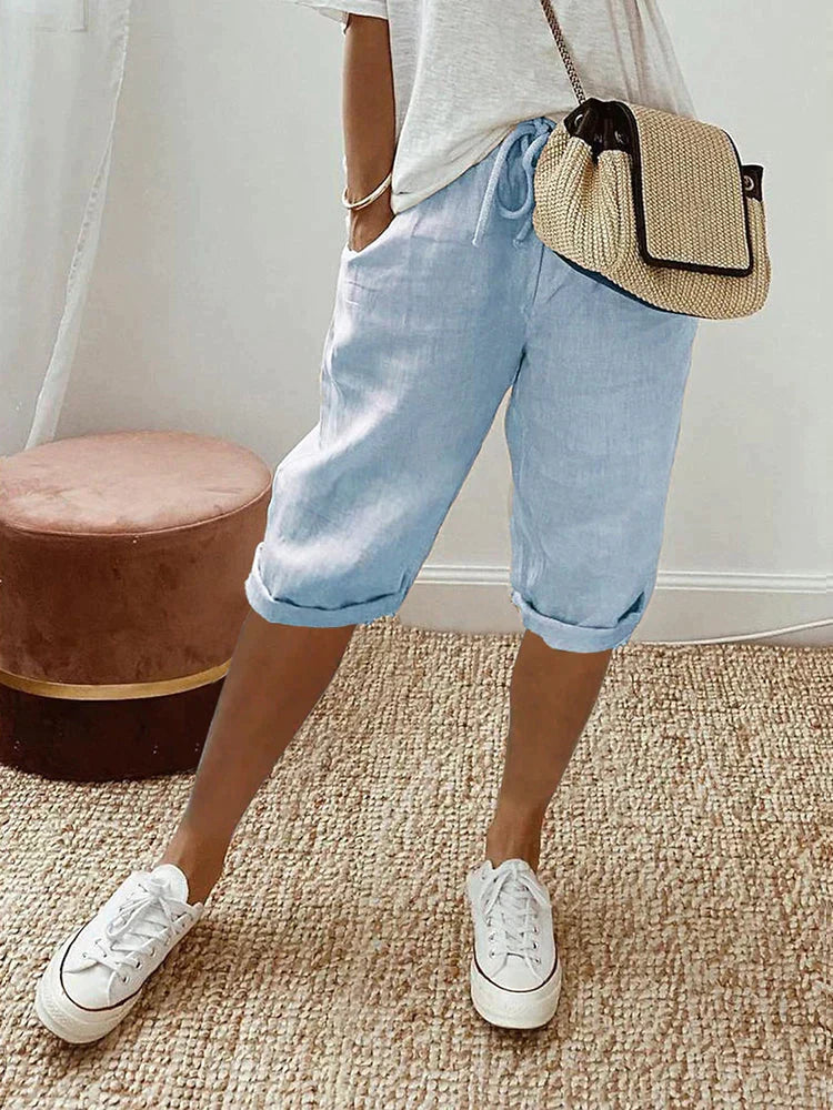 Light blue comfort shorts paired with a white top and casual sneakers, set in a cozy indoor room with woven rug and stool.