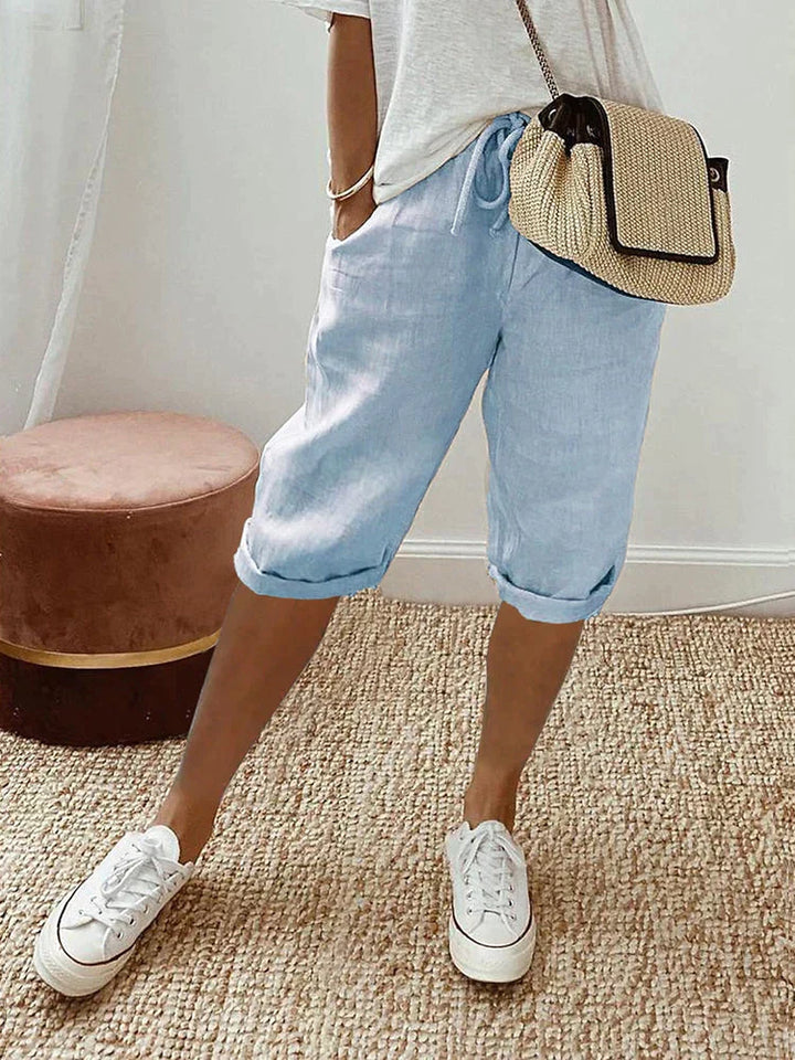 Light blue comfort shorts paired with a white top and casual sneakers, set in a cozy indoor room with woven rug and stool.