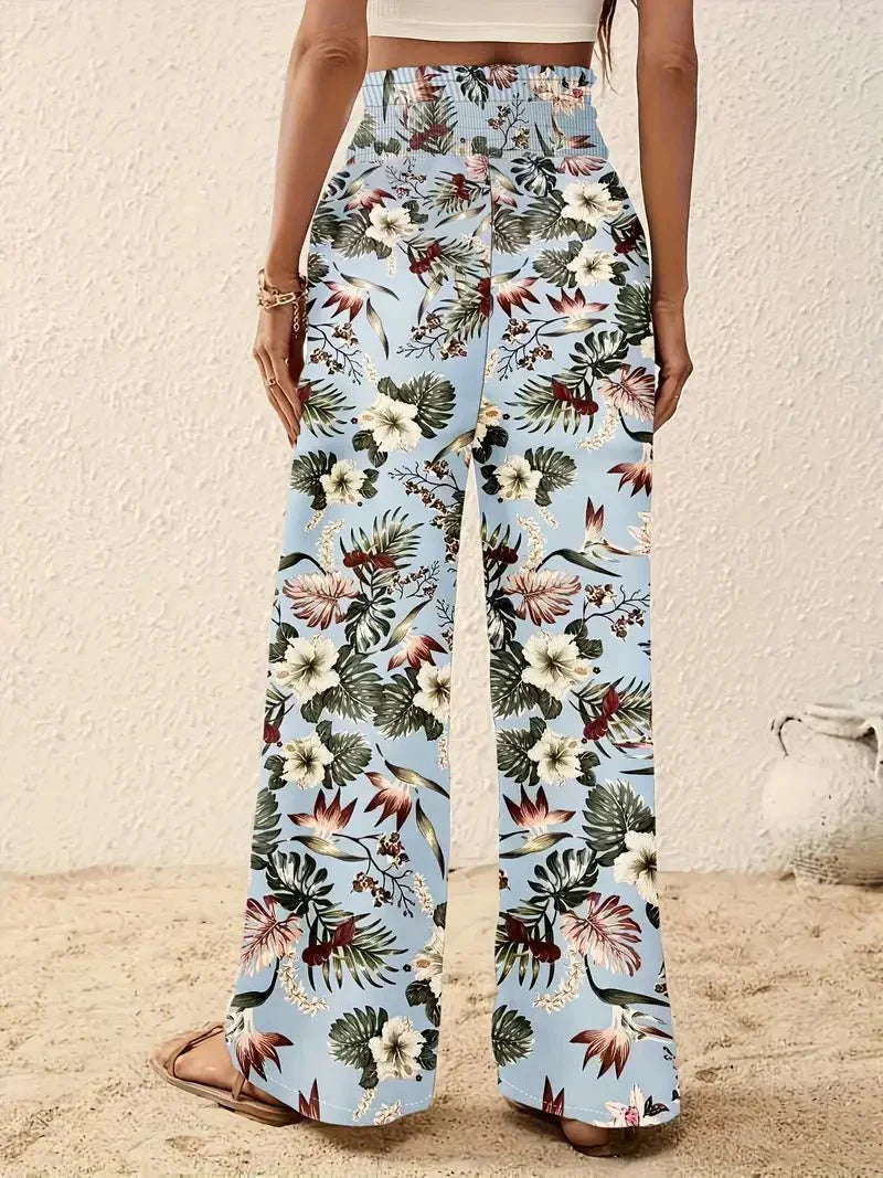 Back view of light blue floral trousers with red and green designs in a sandy, beach-like setting.