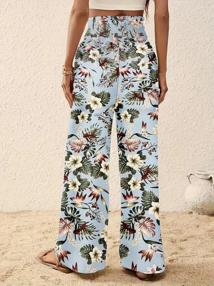 Back view of light blue floral trousers with red and green designs in a sandy, beach-like setting.