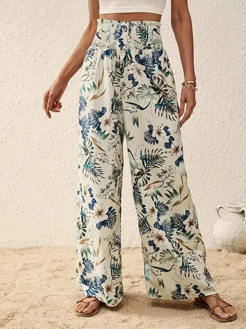 Close-up of elegant floral trousers with blue and green tones against a textured wall and sandy floor.