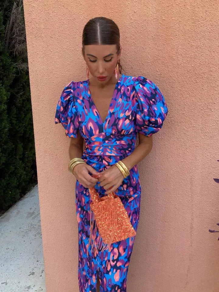 Woman in a vibrant puff sleeve dress with orange purse stands against a peach textured wall with greenery.