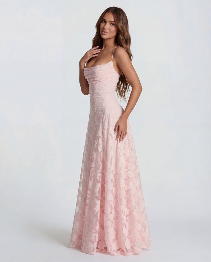 Model posing in a light pink maxi dress with floral details and thin straps, plain studio background.