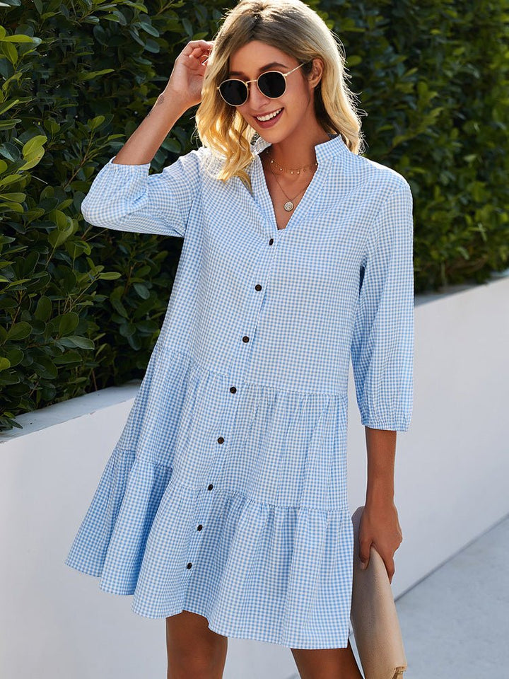 Woman in blue gingham flowy midi dress outdoors with green shrub backdrop.