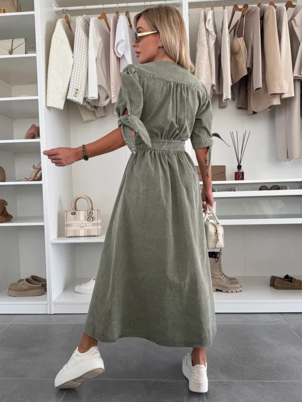 Back view of green denim dress with tied sleeves, worn by a person holding a white bag, in a closet setting.