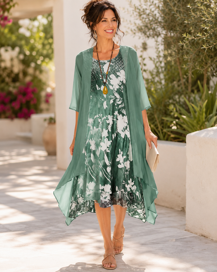 Woman in green floral two-layer dress, walking on a sunlit path lined with blooming plants.
