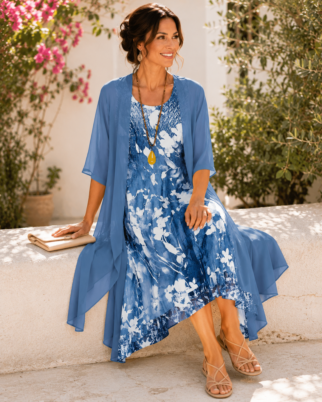 Model sitting on a stone ledge, wearing blue floral two-layer dress, surrounded by colorful flowers.