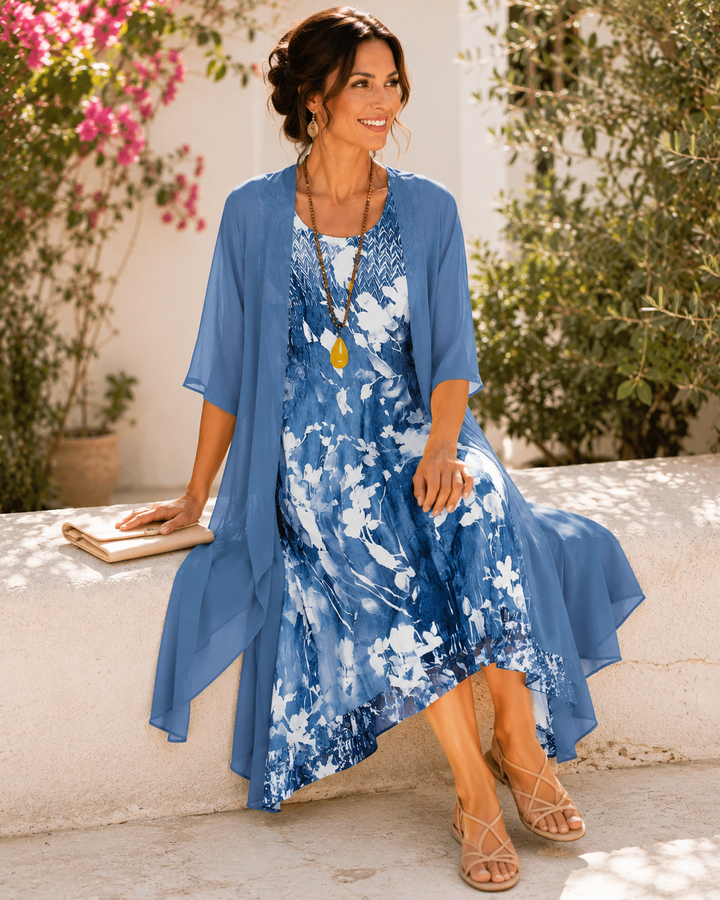Model sitting on a stone ledge, wearing blue floral two-layer dress, surrounded by colorful flowers.
