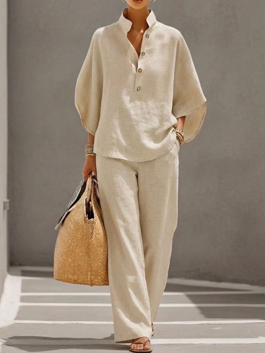 Woman wearing a beige two-piece set with a high collar and buttons, carrying a woven bag, walking in a minimalist urban setting.