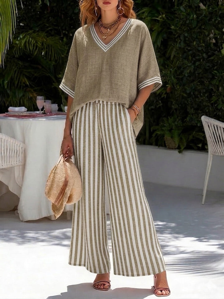 Woman in beige flowy top and striped wide-leg pants, holding a hat, standing in a lush garden.