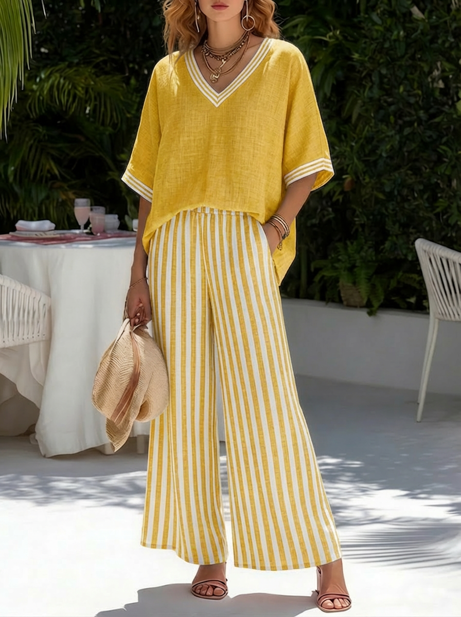 Woman in yellow flowy top and striped wide-leg pants, holding a hat, standing in a lush garden.