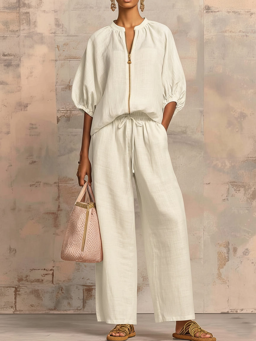 Person in a cream two-piece set with wide pants and puffed sleeves, holding a stylish bag against a textured wall.