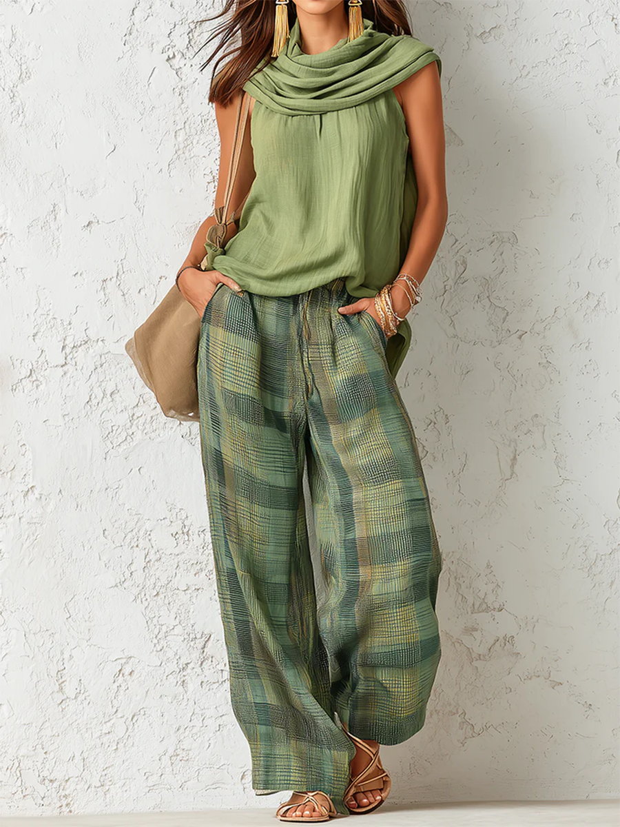Woman in a relaxed green top with a draped neckline and green plaid wide-leg pants, standing against a textured white wall.