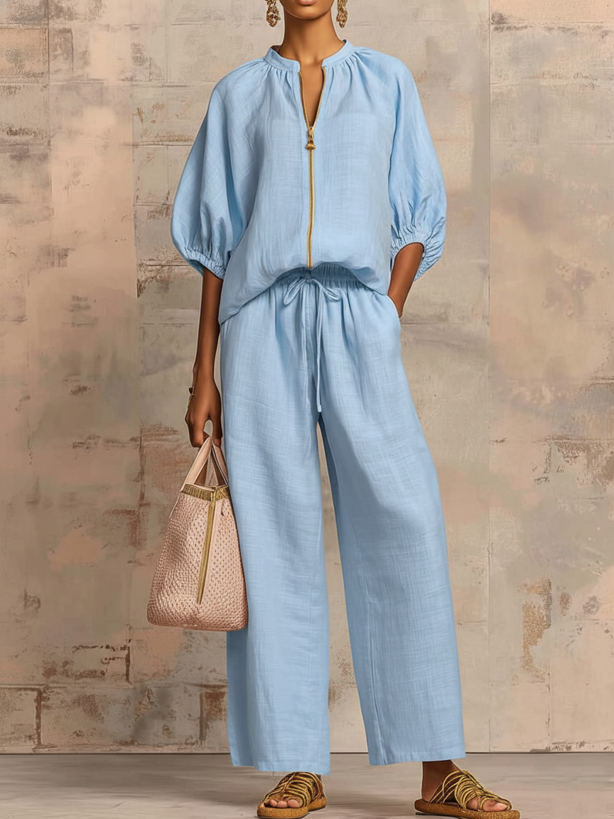 Person in a blue two-piece set with wide pants and puffed sleeves, holding a chic bag against a textured wall.
