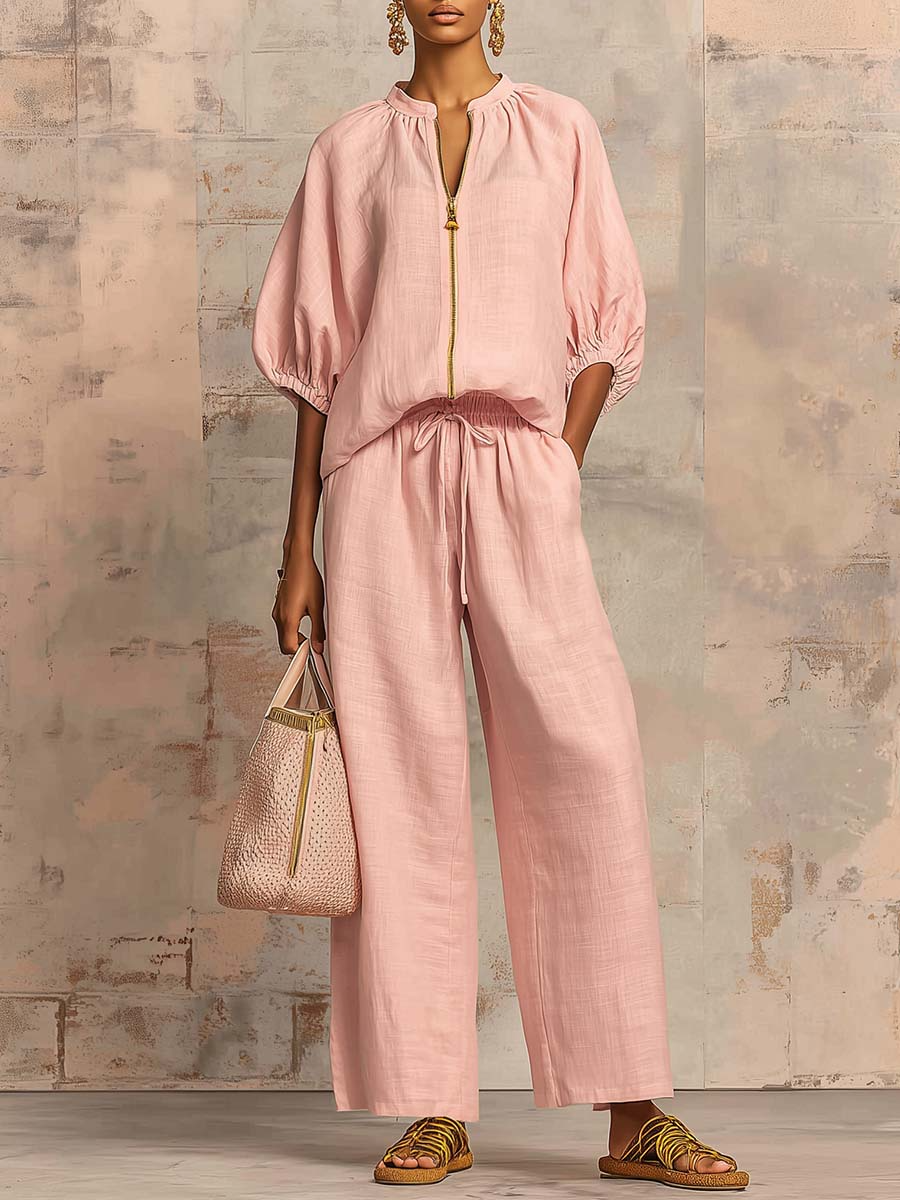 Person wearing a pink two-piece outfit with wide pants and puffed sleeves, holding a stylish bag and standing against a textured wall.