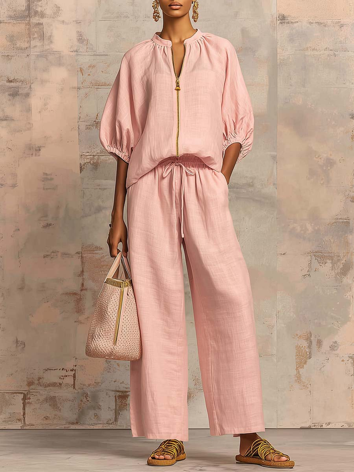 Person wearing a pink two-piece outfit with wide pants and puffed sleeves, holding a stylish bag and standing against a textured wall.