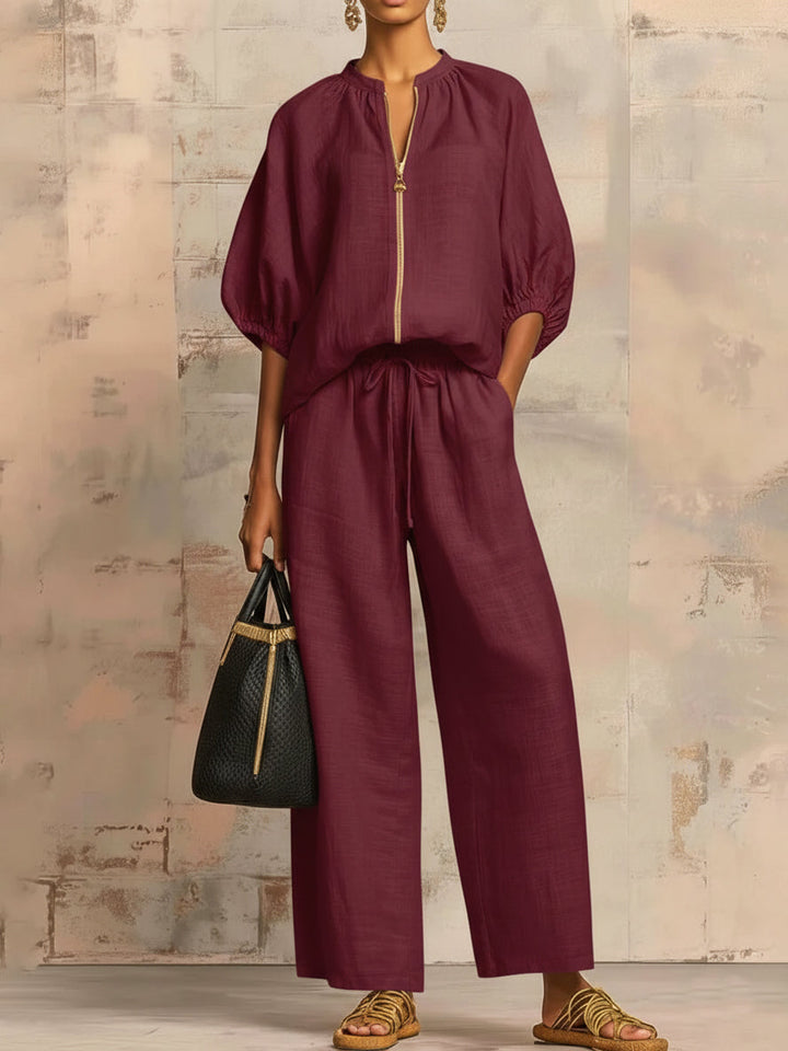Person wearing a burgundy two-piece outfit with wide pants and puffed sleeves, holding a black bag against a textured wall.