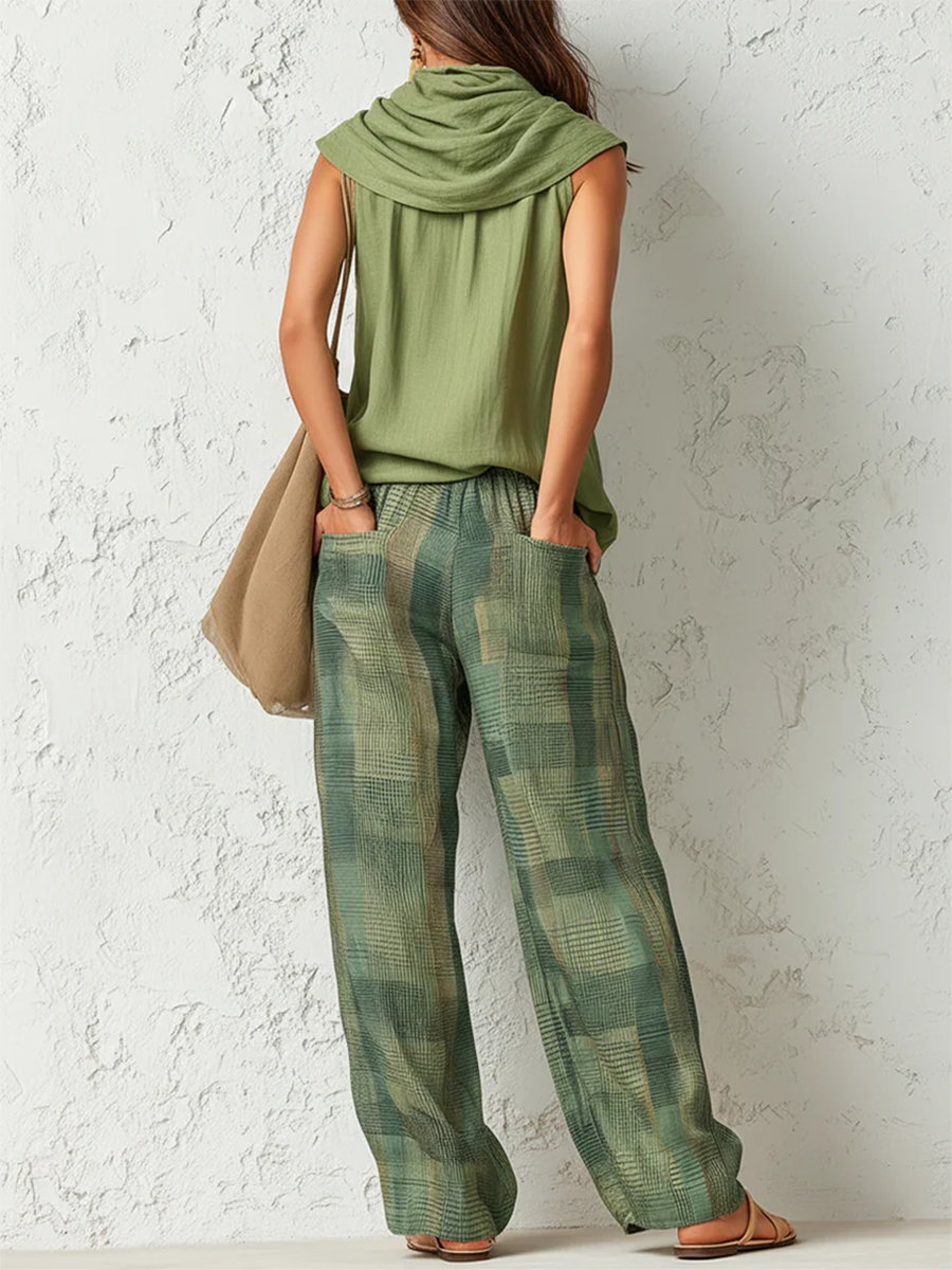Back view of woman wearing a green draped top and plaid wide-leg pants, hands in pockets, against a textured white wall.