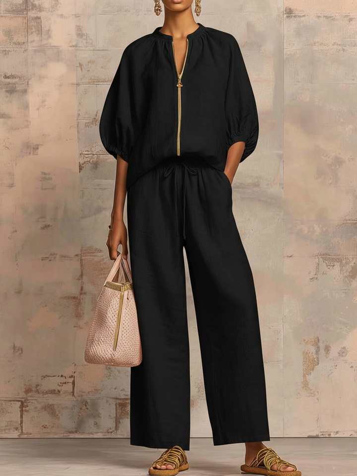 Person wearing a black two-piece outfit with wide pants and puffed sleeves, holding a chic bag against a textured wall.