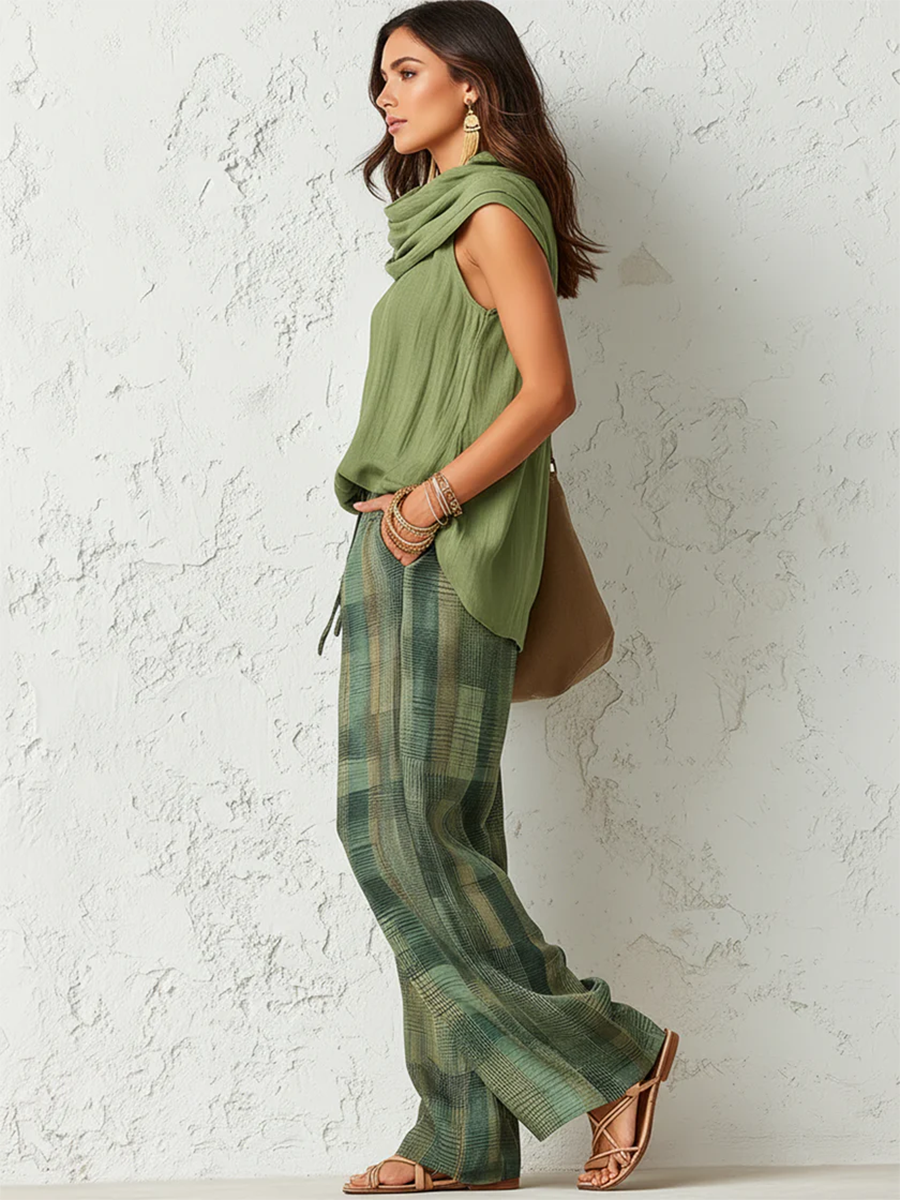 Side view of woman in a green top with draped neckline and plaid pants, standing by a white textured wall.