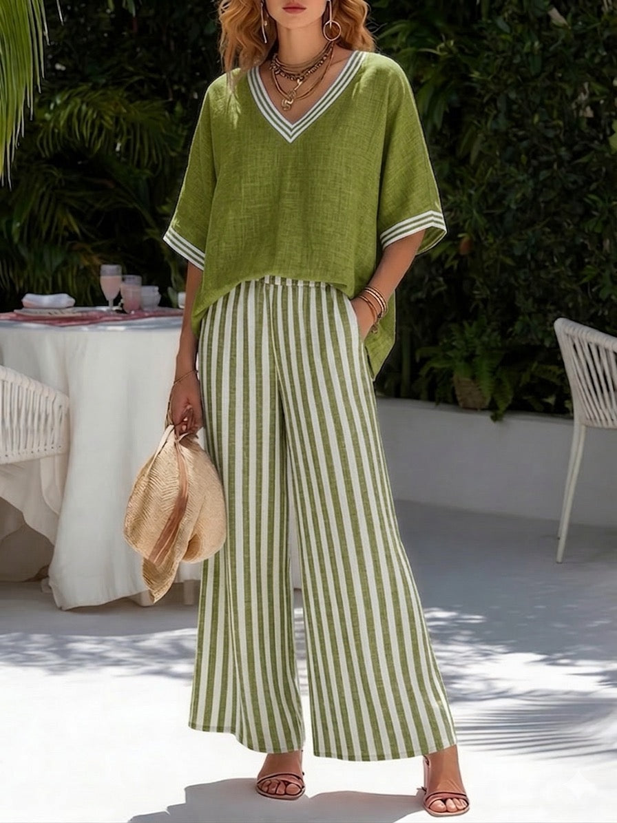Woman in green flowy top and striped wide-leg pants, holding a hat, standing in a lush garden.