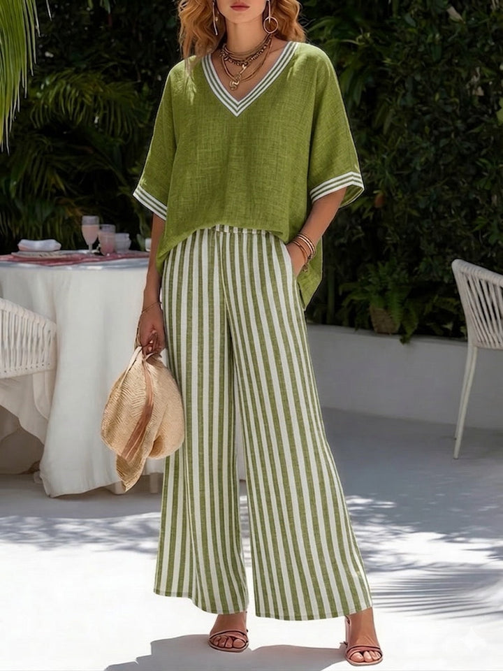 Woman in green flowy top and striped wide-leg pants, holding a hat, standing in a lush garden.
