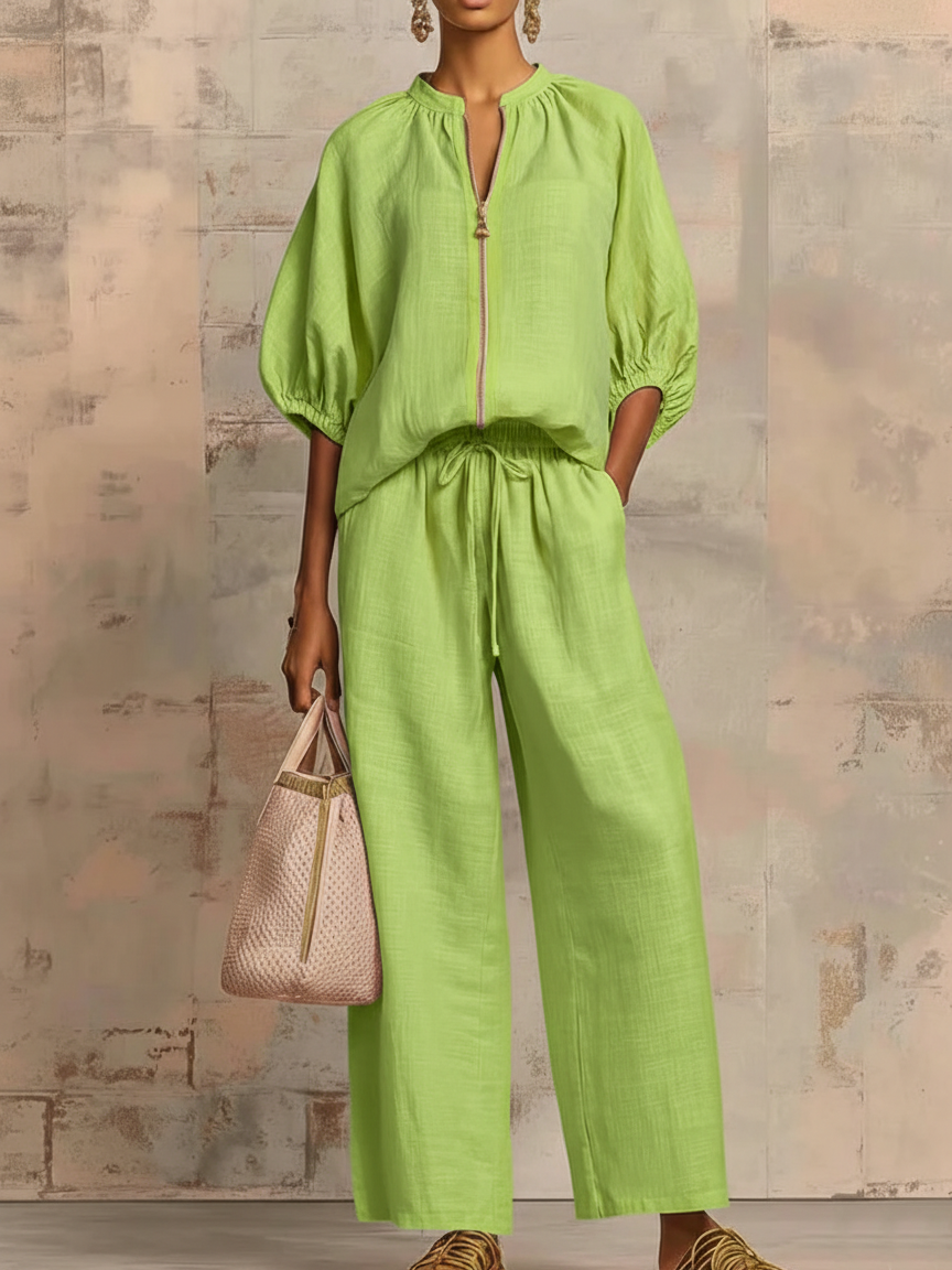 Person in a green two-piece set with wide pants and puffed sleeves, holding a stylish bag against a textured wall.