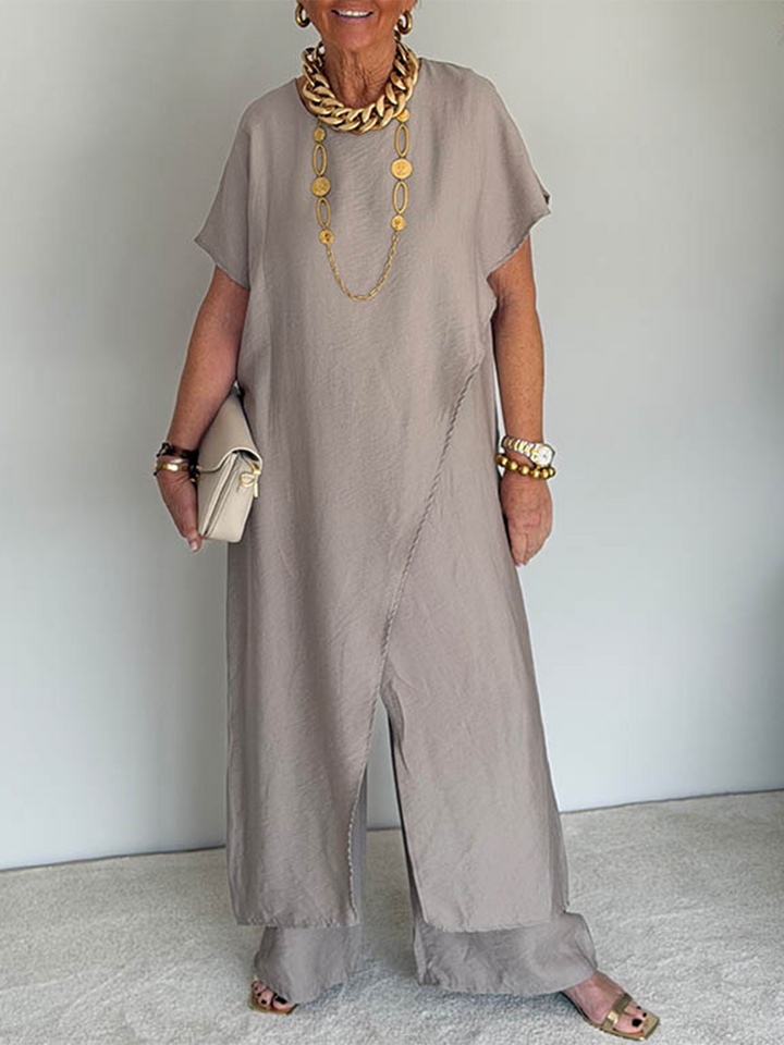 Person wearing a taupe flowing set with short sleeves and wide-leg pants, accessorized with gold jewelry and holding a beige clutch in a studio setting.