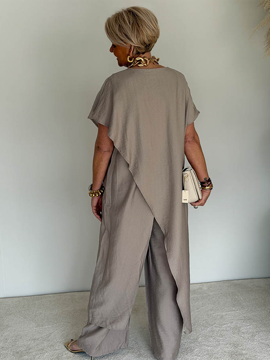 Back view of a person wearing a taupe flowing set with an overlapping design, accessorized with gold jewelry and holding a beige clutch in a studio setting.