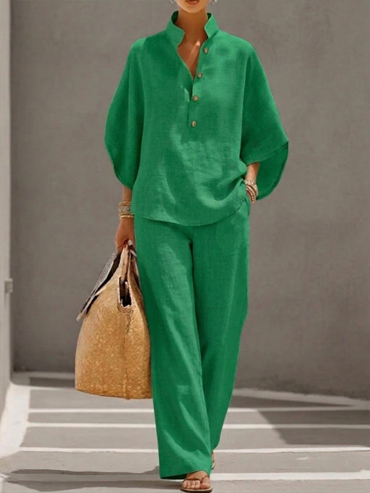 Woman wearing a green two-piece set with a high collar and buttons, carrying a woven bag, walking in a minimalist urban setting.