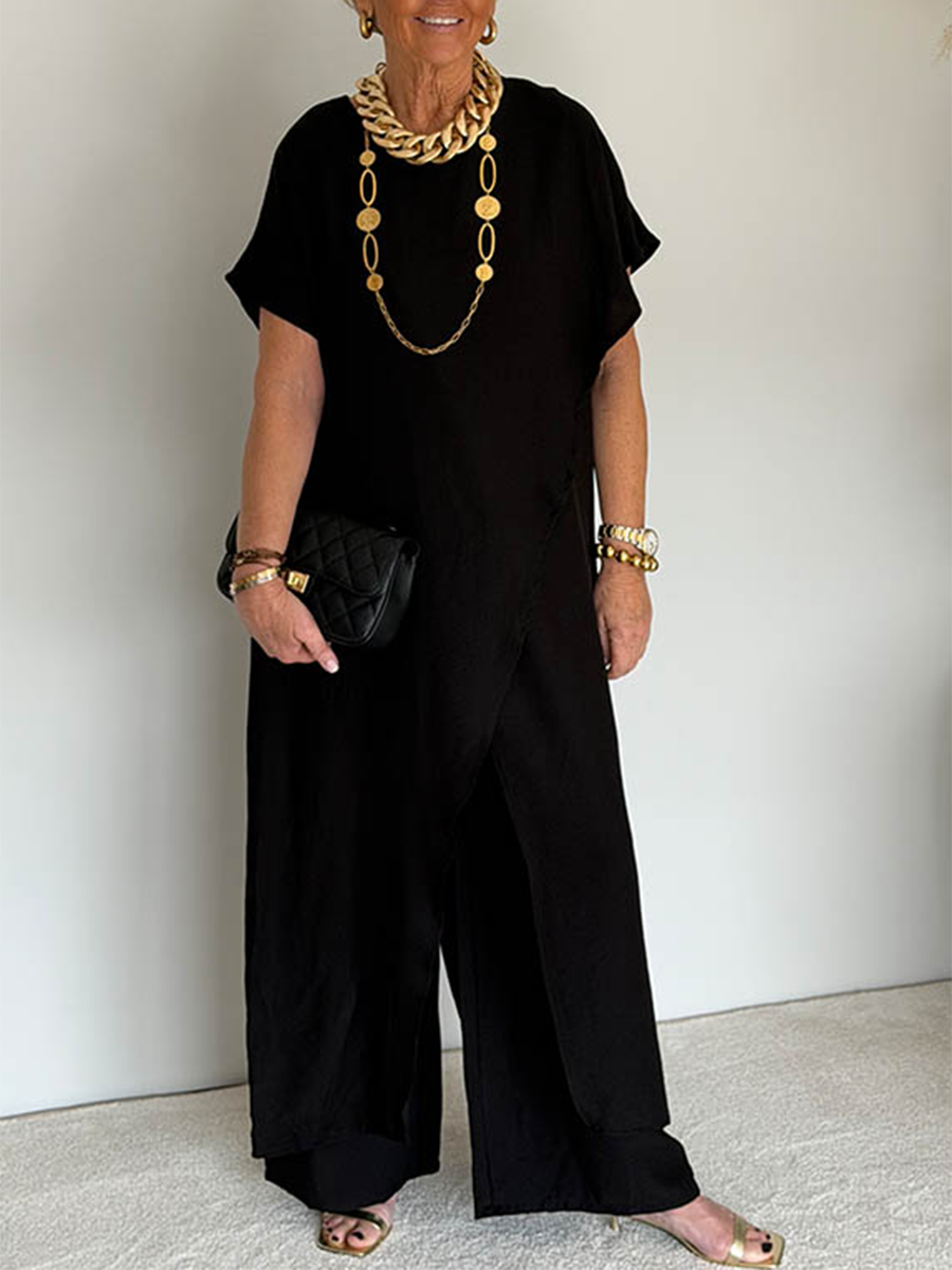 Person wearing a black flowing set with short sleeves and wide-leg pants, accessorized with gold jewelry and holding a black clutch in a studio setting.