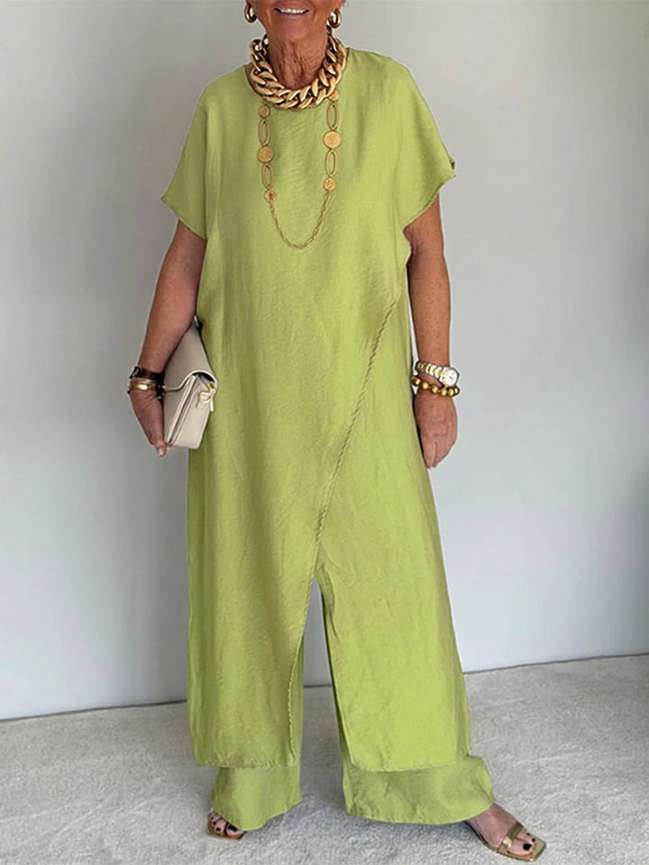 Person wearing a light green flowing set with short sleeves and wide-leg pants, accessorized with gold jewelry and holding a beige clutch in a studio setting.